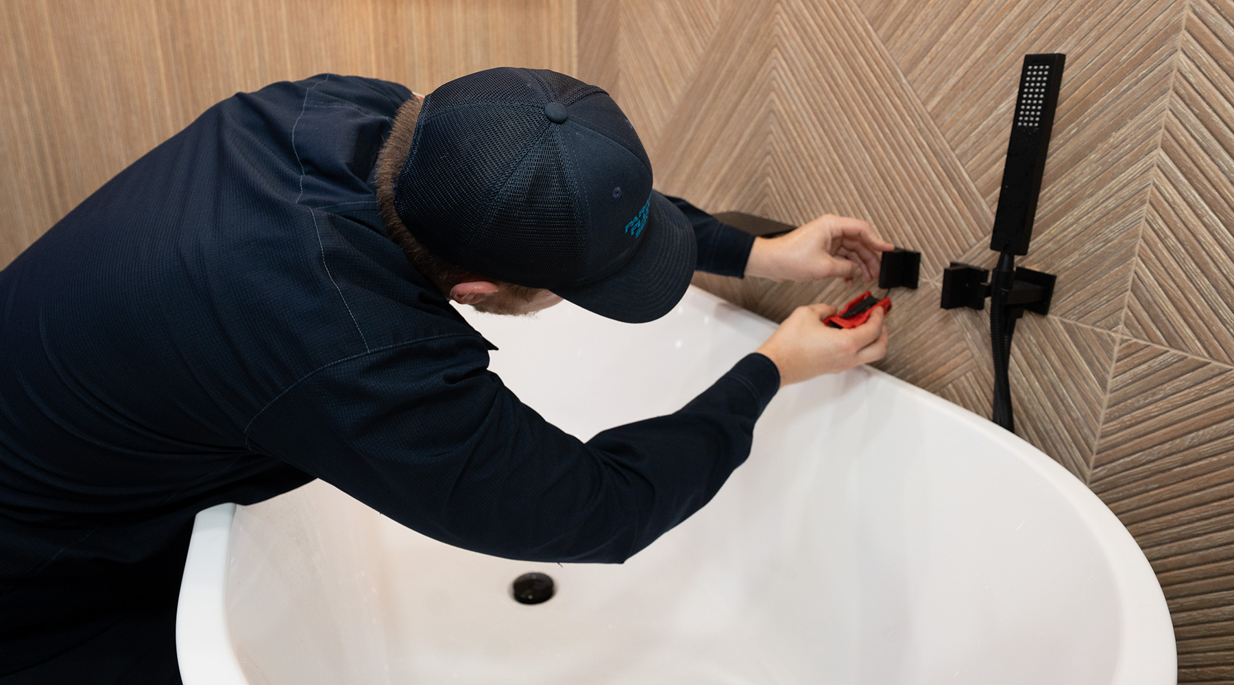 Paramount plumber installs a modern black faucet on a textured wall above a white bathtub, holding a red tool for adjustments.