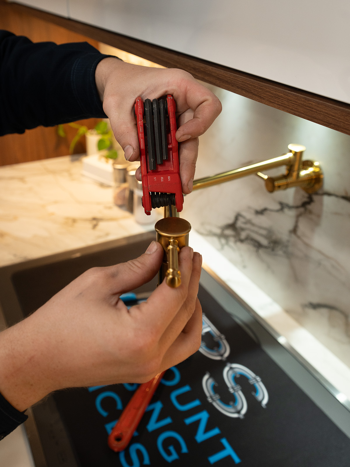 Paramount plumber uses a red tool to install a brass fixture in a stylish kitchen with marble countertops and a modern sink design.
