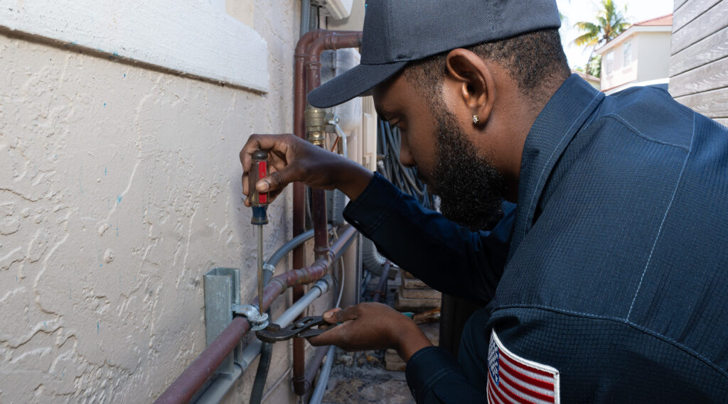 Paramount plumber using a screwdriver and pliers to repair plumbing pipes on an exterior wall