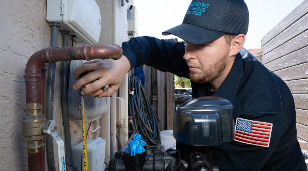 Paramount plumber measures a pipe connection with a tape measure, working on plumbing near a wall with multiple pipes and a water valve.