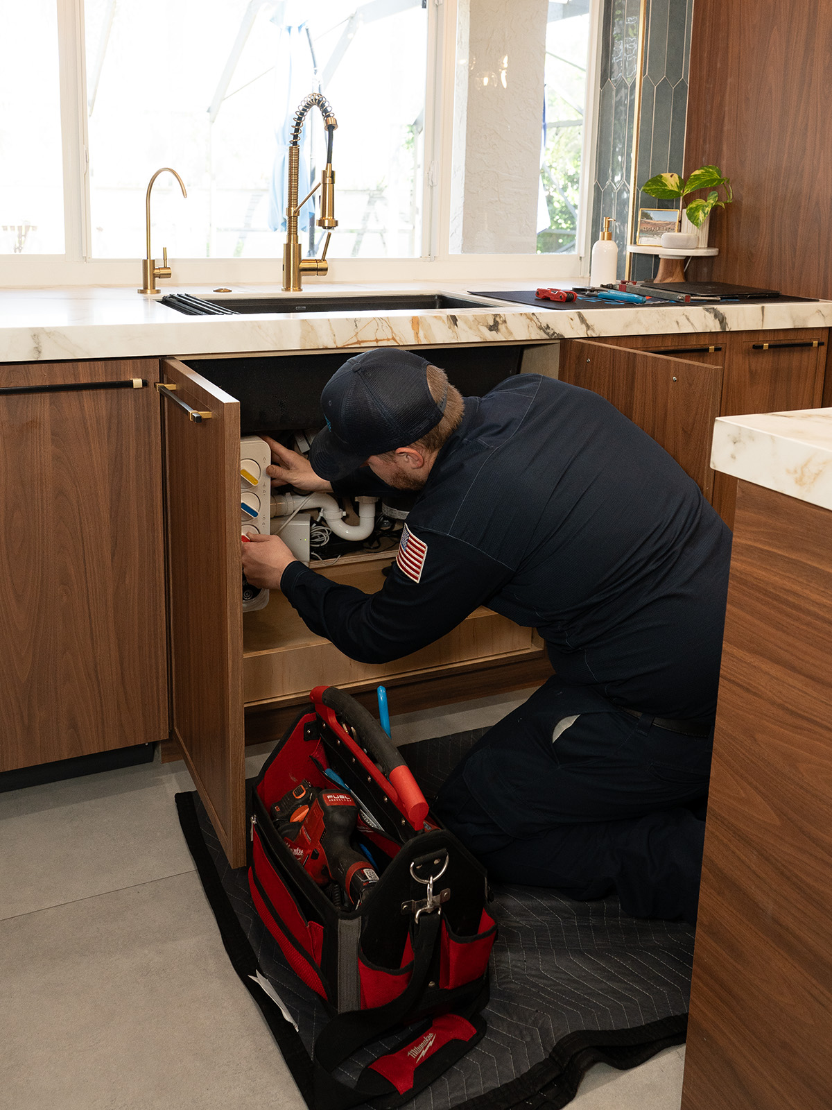 A plumber kneels beside an open kitchen cabinet, working on water filtration system under sink