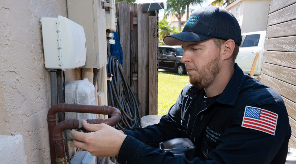 Paramount plumber in a dark uniform works on plumbing pipes attached to a wall, with tools and equipment visible in the background.