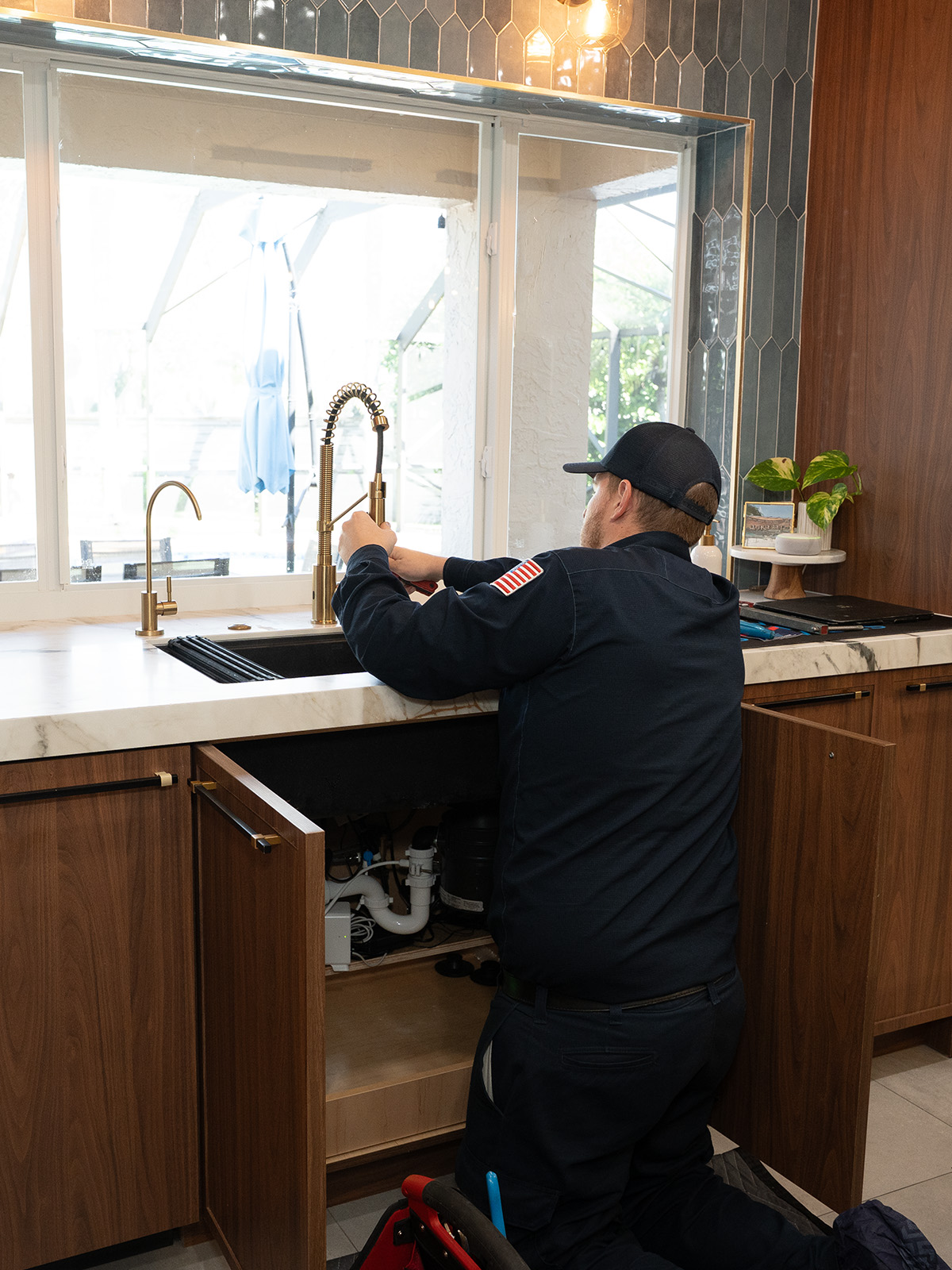 A technician kneels under a kitchen sink, repairing plumbing while the modern faucet and bright window are visible above him.