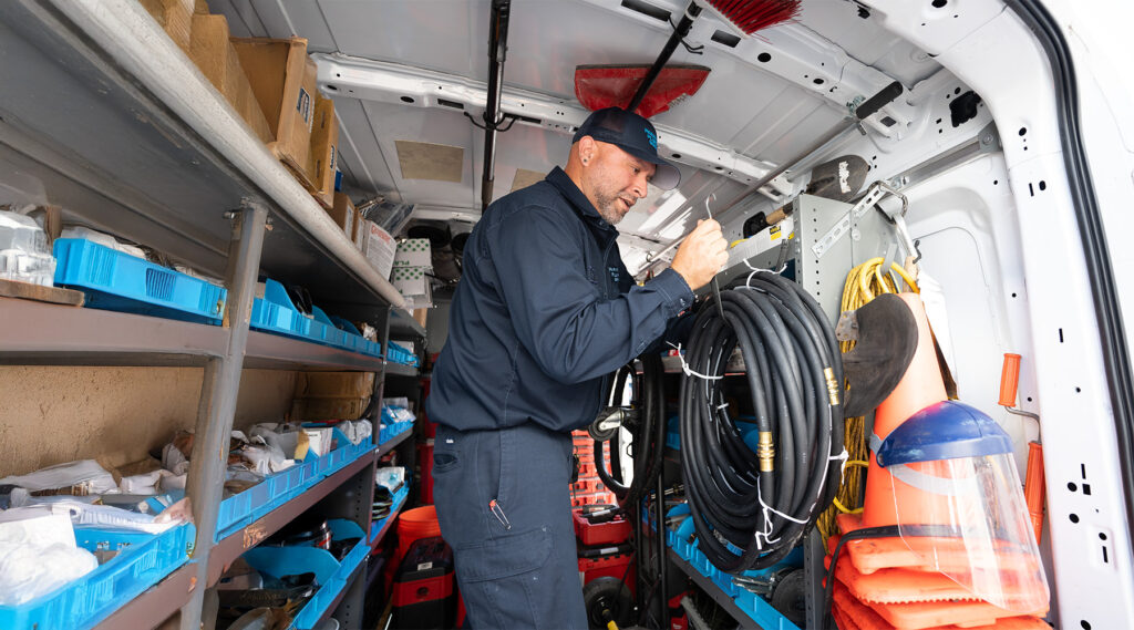 Paramount plumber organizes tools and equipment in a work van, surrounded by shelves filled with blue bins, cables, and safety gear.